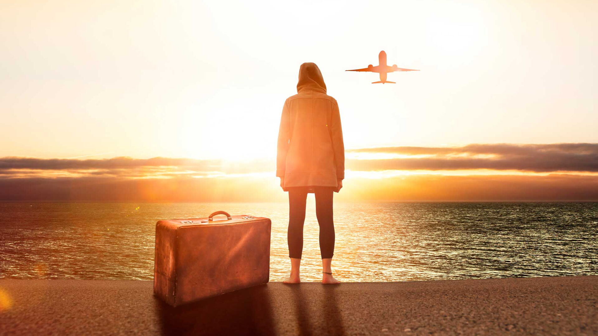 Eine Person steht barfuß mit Koffer am Meer und schaut einem Flugzeug im Sonnenuntergang nach.