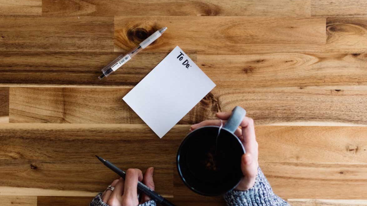 Eine Hand hält einen Stift, die andere eine Tasse Tee, daneben liegt ein leeres To-do-Blatt auf dem Tisch.