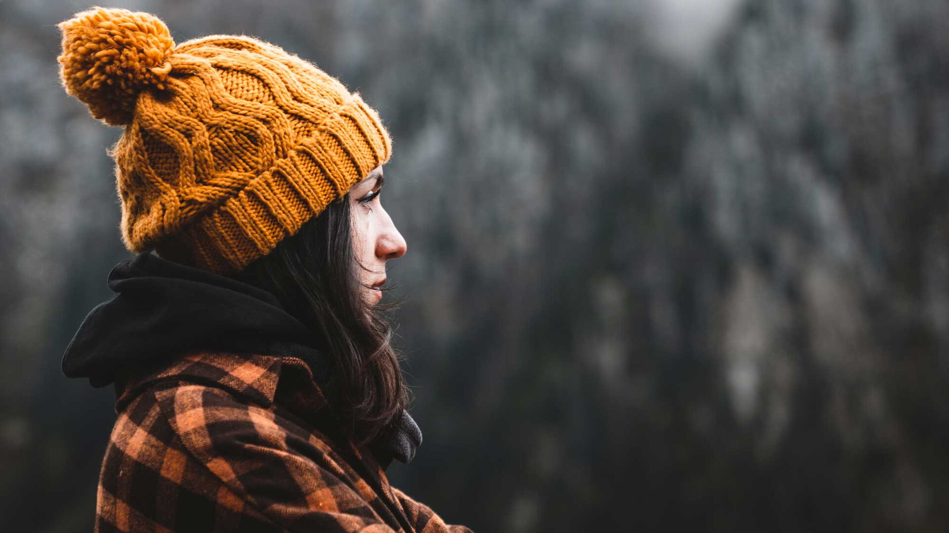 Seitenansicht einer nachdenklichen jungen Frau, die ein kariertes Hemd und eine gelbe Wintermütze trägt und bei kaltem Wetter die Aussicht betrachtet.