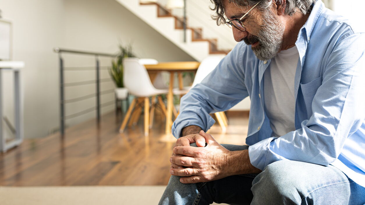 Reifer Mann mit Knieschmerzen sitzt auf dem Sofa im Wohnzimmer zu Hause.